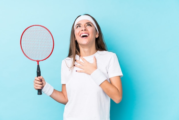 Jovem mulher caucasiana jogando badminton isolado ri alto, mantendo a mão no peito.