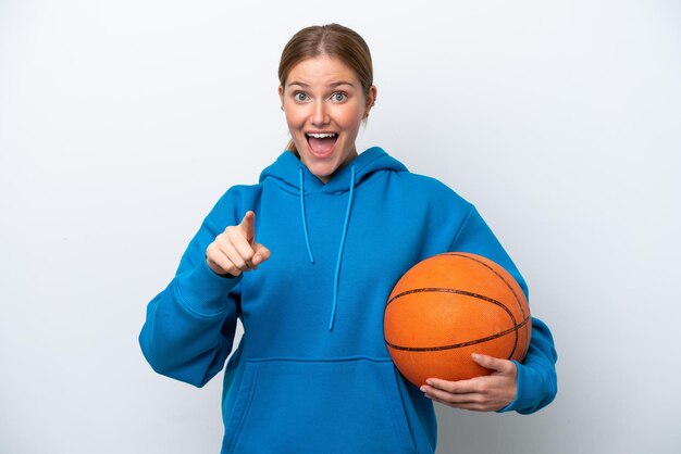 Jovem mulher caucasiana jogando basquete isolada no fundo branco surpresa e apontando para a frente