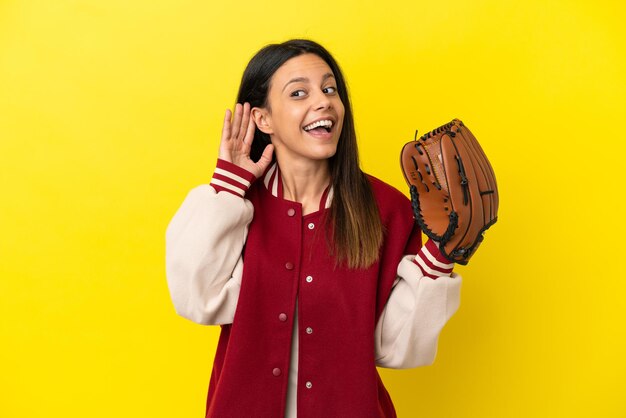 Jovem mulher caucasiana jogando beisebol isolada em um fundo amarelo, ouvindo algo colocando a mão na orelha