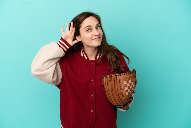 Jovem mulher caucasiana jogando beisebol isolada em um fundo azul, ouvindo algo colocando a mão na orelha