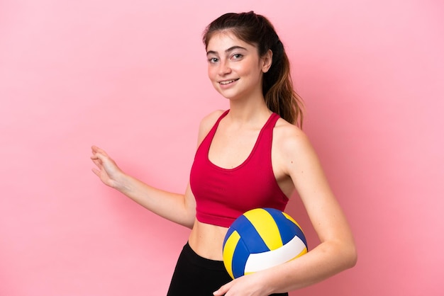 Jovem mulher caucasiana jogando vôlei isolada em fundo rosa, estendendo as mãos para o lado para convidar para vir