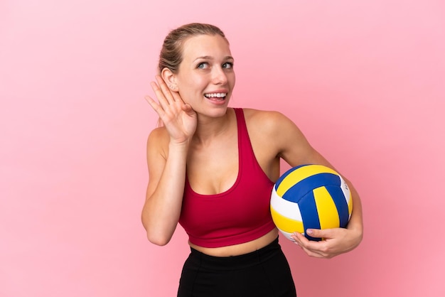 Jovem mulher caucasiana jogando vôlei isolada em um fundo rosa, ouvindo algo colocando a mão na orelha