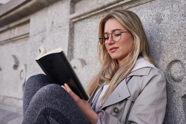 Jovem mulher caucasiana lendo livro no parque com prédio antigo