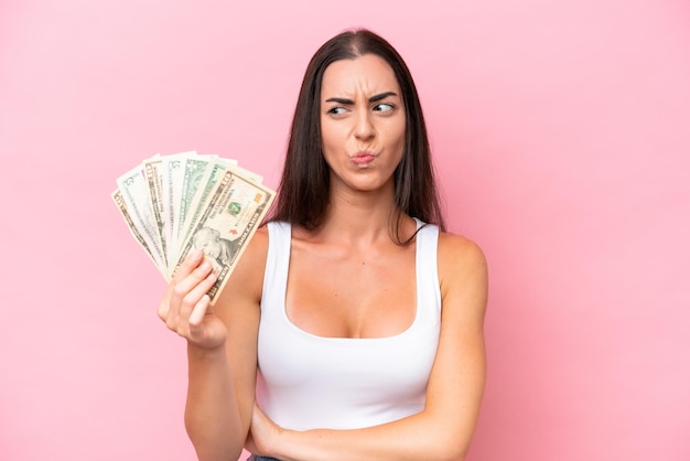 Jovem mulher caucasiana levando muito dinheiro isolado no fundo rosa com expressão triste