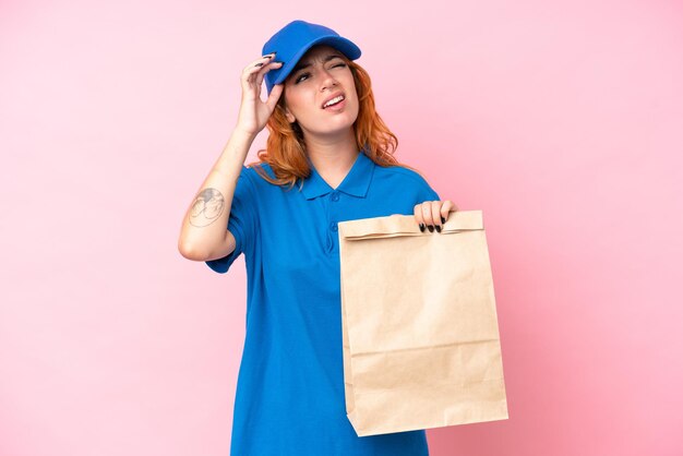 Jovem mulher caucasiana levando um saco de comida para viagem isolada em fundo rosa com dúvidas e pensando