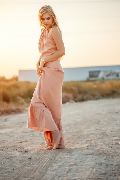 Jovem mulher caucasiana linda num vestido rosa caminha na areia com os pés descalços durante o pôr do sol