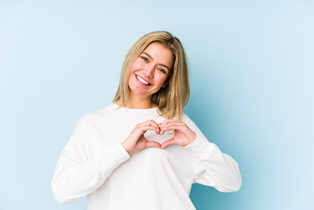 Jovem mulher caucasiana loira isolada sorrindo e mostrando uma forma de coração com as mãos.