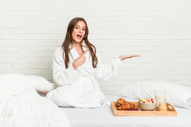 Jovem mulher caucasiana na cama animada, segurando um espaço de cópia na palma da mão.