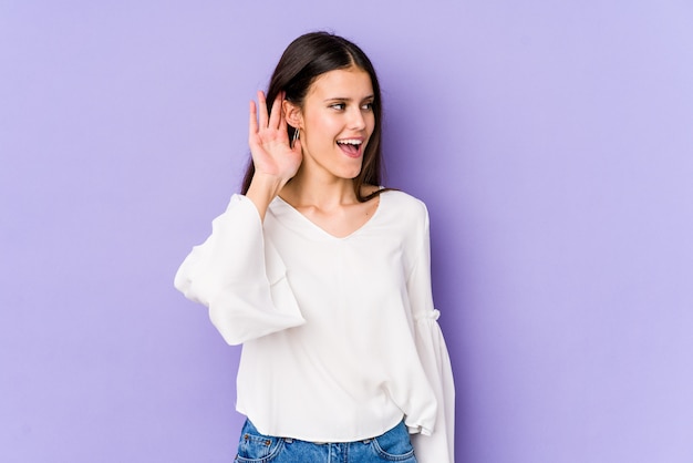 Jovem mulher caucasiana na parede roxa, tentando ouvir uma fofoca.