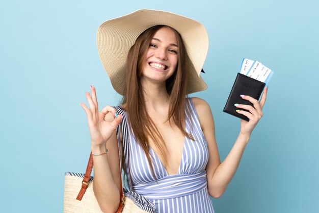Jovem mulher caucasiana nas férias de verão segurando o passaporte isolado em um fundo azul e mostrando o sinal de ok com os dedos