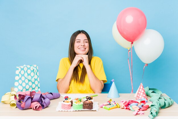 Jovem mulher caucasiana, organizando um aniversário mantém as mãos sob o queixo, está olhando alegremente de lado.