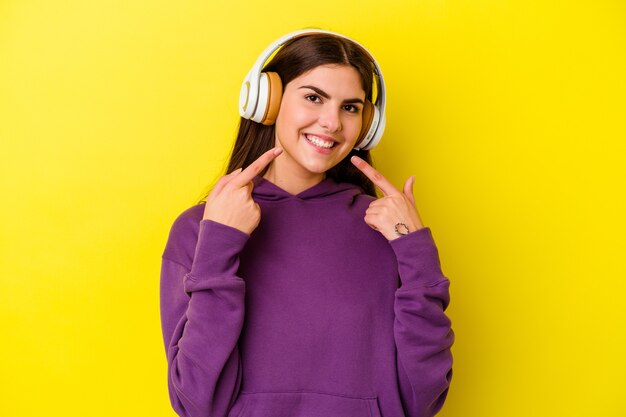 Jovem mulher caucasiana, ouvindo música com fones de ouvido isolados em sorrisos de fundo rosa, apontando os dedos na boca.