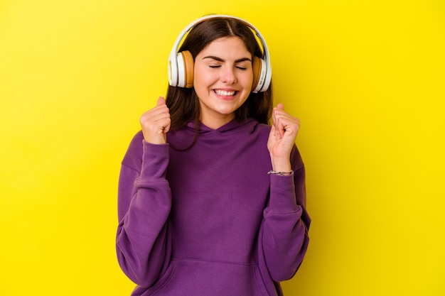 Jovem mulher caucasiana ouvindo música com fones de ouvido isolados na parede rosa levantando o punho, sentindo-se feliz e bem-sucedida