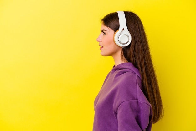 Jovem mulher caucasiana, ouvindo música com fones de ouvido isolados na parede rosa, olhando para a esquerda, pose de lado.
