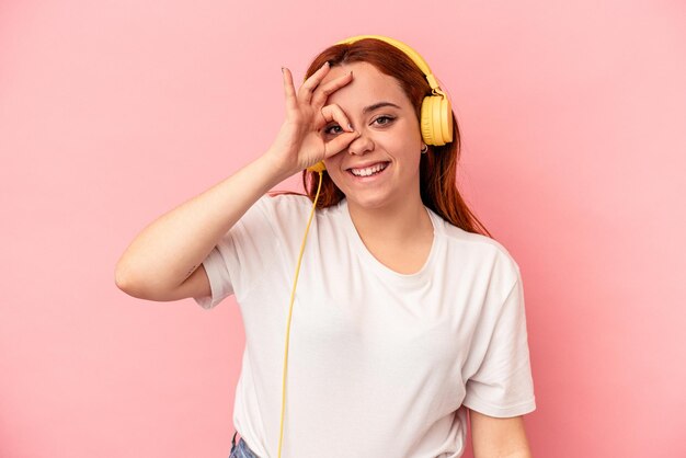 Jovem mulher caucasiana, ouvindo música isolada no fundo rosa, animada em manter o gesto ok no olho.