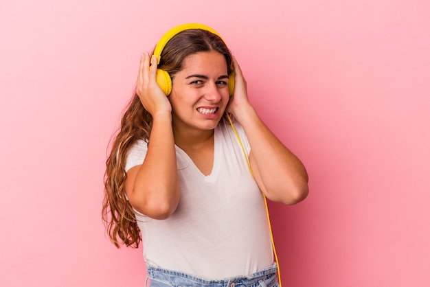 Jovem mulher caucasiana, ouvindo música isolada no fundo rosa, cobrindo as orelhas com as mãos.