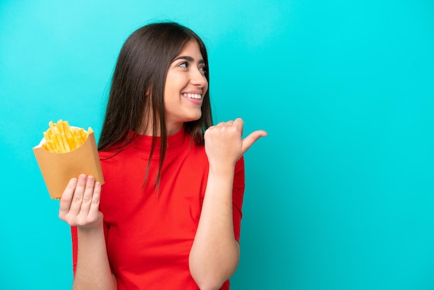 Jovem mulher caucasiana pegando batatas fritas isoladas em fundo azul, apontando para o lado para apresentar um produto