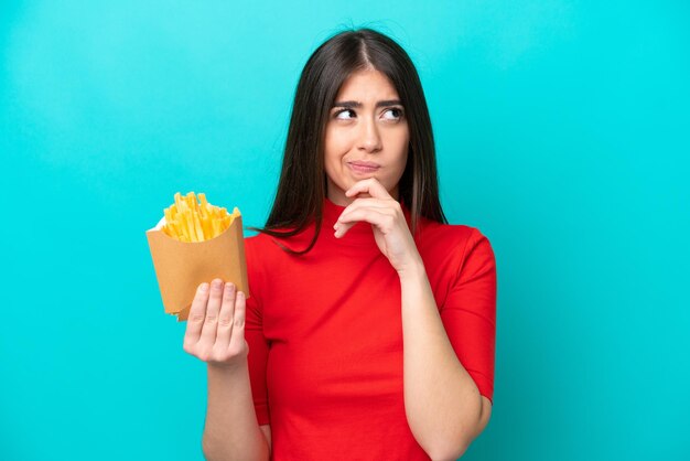 Jovem mulher caucasiana pegando batatas fritas isoladas em fundo azul com dúvidas e pensando