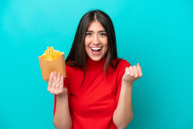 Jovem mulher caucasiana pegando batatas fritas isoladas em fundo azul comemorando uma vitória na posição de vencedor