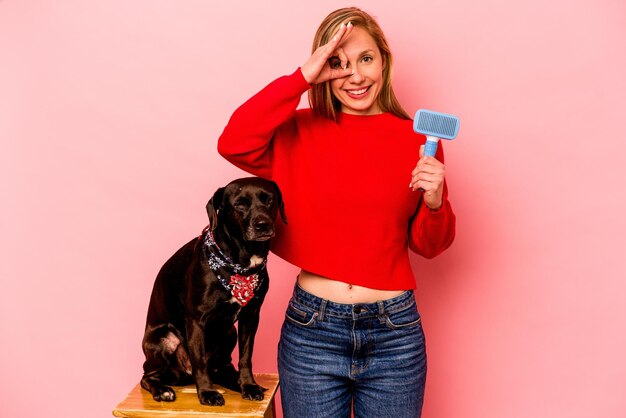 Jovem mulher caucasiana penteando o cachorro isolado no fundo rosa animado mantendo o gesto okey no olho