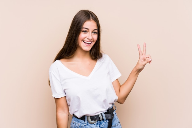 Jovem mulher caucasiana posando isolada, mostrando sinal de vitória e sorrindo amplamente.