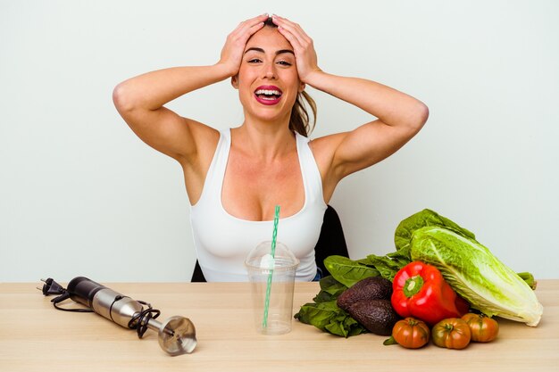 Jovem mulher caucasiana, preparando um smoothie saudável com legumes, ri mantendo as mãos na cabeça com alegria. Conceito de felicidade.