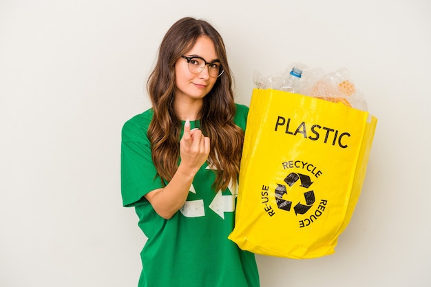 Jovem mulher caucasiana, reciclando um cheio de plástico isolado no fundo branco, apontando com o dedo para você, como se fosse um convite para se aproximar.