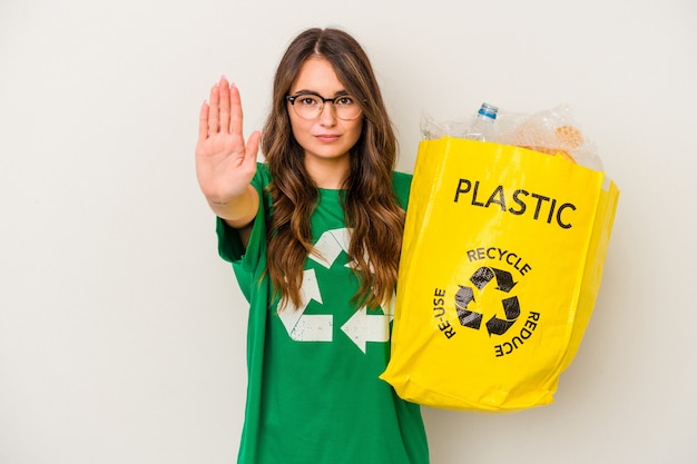 Jovem mulher caucasiana, reciclando um cheio de plástico isolado no fundo branco, de pé com a mão estendida, mostrando o sinal de pare, impedindo-o.