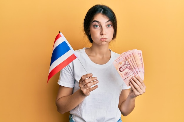 Jovem mulher caucasiana segurando a bandeira da tailândia e notas de baht bufando as bochechas com cara engraçada. boca inflada com ar, pegando ar.