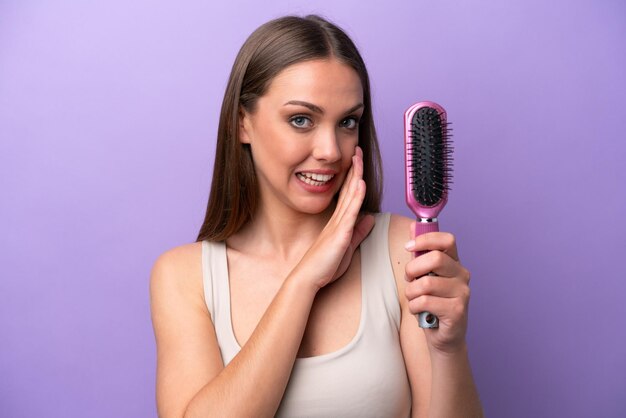 Jovem mulher caucasiana segurando a escova de cabelo isolada no fundo roxo sussurrando algo