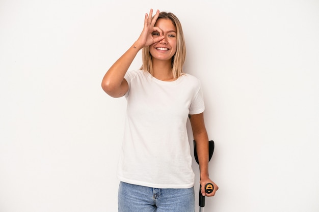 Jovem mulher caucasiana, segurando a muleta isolada no fundo branco, animado, mantendo o gesto ok no olho.