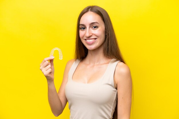 Jovem mulher caucasiana segurando aparelho invisível isolado em fundo amarelo sorrindo muito