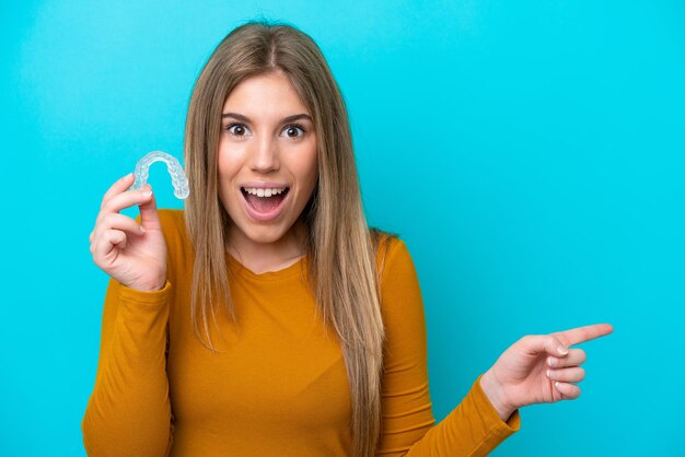 Foto jovem mulher caucasiana segurando aparelho invisível isolado em fundo azul surpreso e apontando o dedo para o lado