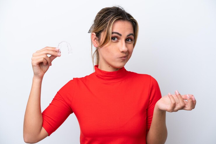 Foto jovem mulher caucasiana segurando aparelho invisível no fundo branco, fazendo gesto de dúvidas enquanto levanta os ombros