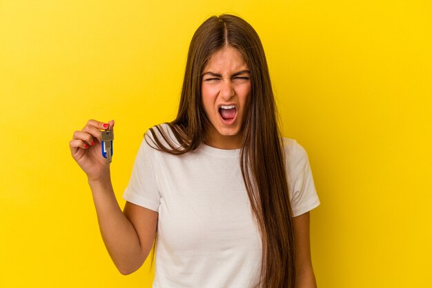 Jovem mulher caucasiana, segurando as chaves de uma casa isoladas em um fundo amarelo, gritando com muita raiva e agressividade.