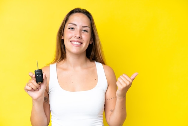 Jovem mulher caucasiana segurando as chaves do carro isoladas em fundo amarelo apontando para o lado para apresentar um produto