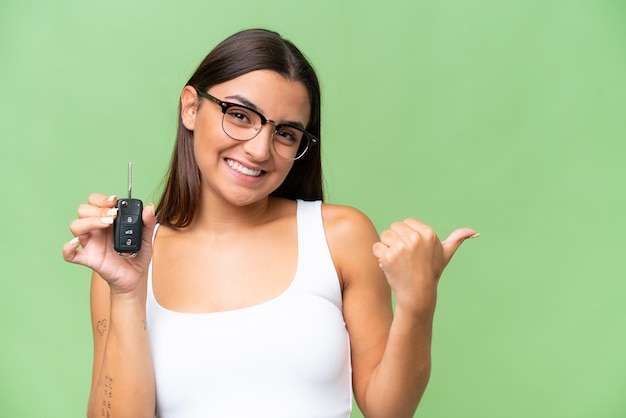 Jovem mulher caucasiana segurando as chaves do carro isoladas no fundo croma verde apontando para o lado para apresentar um produto