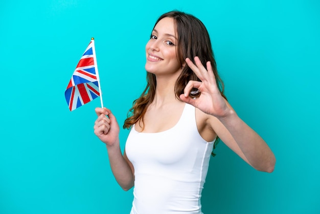 Jovem mulher caucasiana segurando bandeira inglesa isolada em fundo azul, mostrando sinal de ok com os dedos