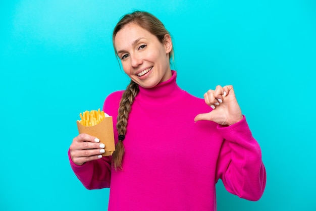 Jovem mulher caucasiana segurando batatas fritas isoladas em fundo azul orgulhosa e satisfeita