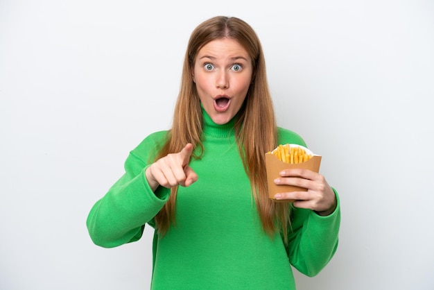 Jovem mulher caucasiana segurando batatas fritas isoladas no fundo branco surpreso e apontando para a frente