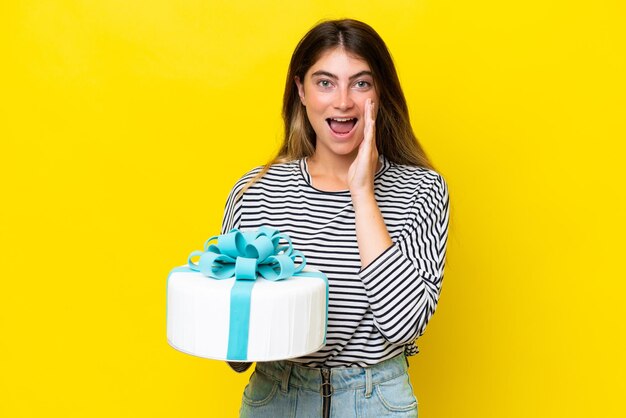 Jovem mulher caucasiana segurando bolo de aniversário isolado em fundo amarelo gritando com a boca aberta