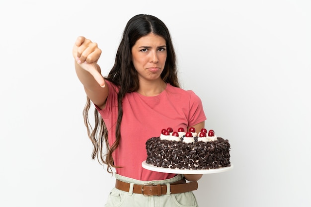 Jovem mulher caucasiana segurando bolo de aniversário isolado no fundo branco, mostrando o polegar para baixo com expressão negativa