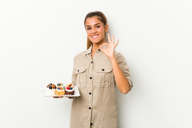 Jovem mulher caucasiana segurando bolos doces alegre e confiante, mostrando um gesto de ok.