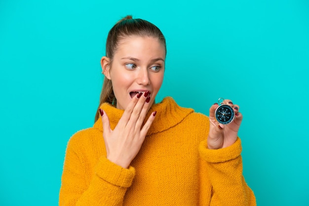 Jovem mulher caucasiana segurando bússola isolada em fundo azul com surpresa e expressão facial chocada