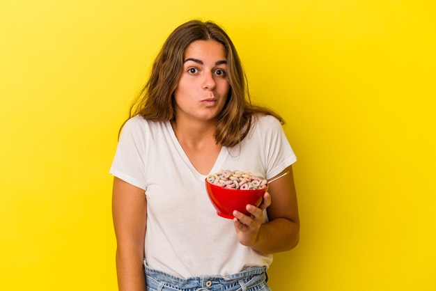 Jovem mulher caucasiana segurando cereais isolados em fundo amarelo encolhe os ombros e abre os olhos confusos.