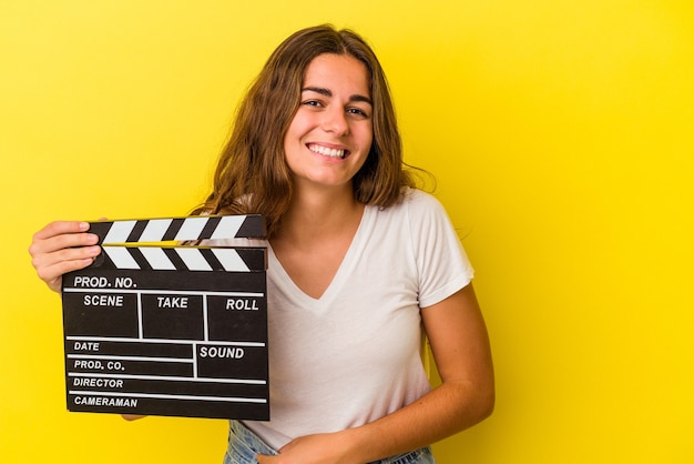 Jovem mulher caucasiana, segurando claquete isolada em fundo amarelo, rindo e se divertindo.