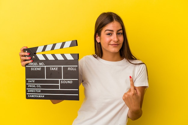 Jovem mulher caucasiana segurando claquete isolada no fundo branco, apontando com o dedo para você como se fosse um convite para se aproximar.