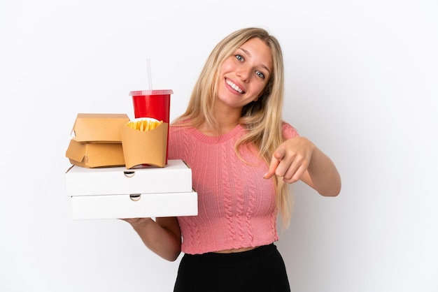 Jovem mulher caucasiana segurando comida gorda isolada em fundo azul, apontando para a frente com expressão feliz
