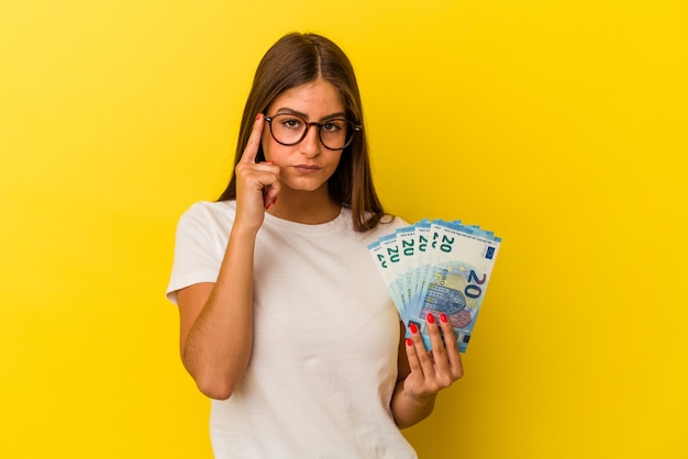 Jovem mulher caucasiana segurando contas isoladas em fundo amarelo, apontando o templo com o dedo, pensando, focada em uma tarefa.