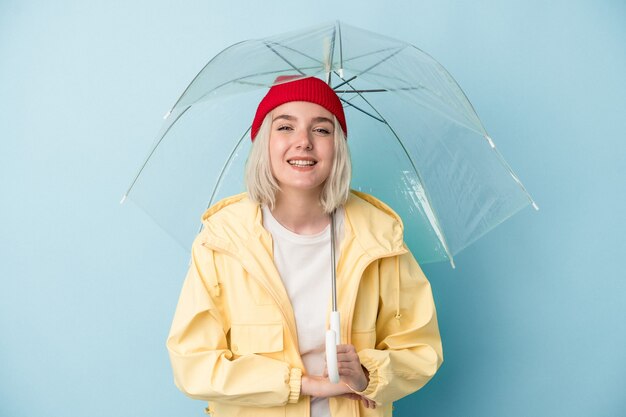 Jovem mulher caucasiana, segurando guarda-chuva isolado em um fundo azul, rindo e se divertindo.